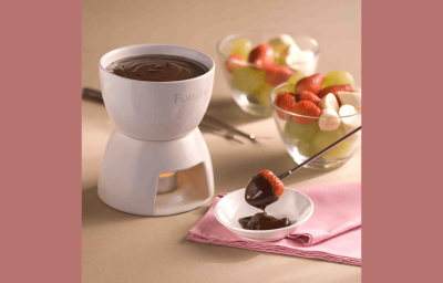 Fotografia de uma panela de fondue de cerâmica branca e aquecida com uma vela abaixo. Ao lado frutas frescas como morango, uva e banana em dois recipientes de vidro e um garfo próprio para fondue com uma morango coberto de chocolate. Fotografia de uma panela de fondue de cerâmica branca e aquecida com uma vela abaixo. Ao lado frutas frescas como morango, uva e banana em dois recipientes de vidro e um garfo próprio para fondue com uma morango coberto de chocolate.