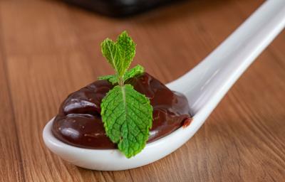 Fotografia de uma colher de servir branca de vidro, com uma porção de brigadeiro com duas folhas de hortelã por cima. A colher está apoiada sobre uma mesa de madeira. Fotografia de uma colher de servir branca de vidro, com uma porção de brigadeiro com duas folhas de hortelã por cima. A colher está apoiada sobre uma mesa de madeira.