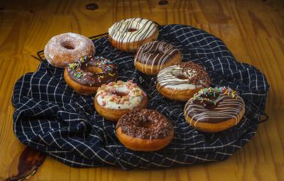 Foto aproximada de uma bancada de madeira com uma grade coberta por um tecido azul quadriculado e sobre ele há oito donuts, alguns decorados com chocolate branco, outros com chocolate ao leite e alguns com confeitos comestíveis Foto aproximada de uma bancada de madeira com uma grade coberta por um tecido azul quadriculado e sobre ele há oito donuts, alguns decorados com chocolate branco, outros com chocolate ao leite e alguns com confeitos comestíveis