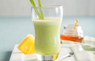 Fotografia em tons de verde em uma mesa de madeira azul clara com um prato retangular branco ao centro com um copo de vidro em cima e o suco de abacaxi, capim-santo e mel dentro dele. Ao fundo, um potinho de vidro com mel. Fotografia em tons de verde em uma mesa de madeira azul clara com um prato retangular branco ao centro com um copo de vidro em cima e o suco de abacaxi, capim-santo e mel dentro dele. Ao fundo, um potinho de vidro com mel.