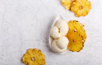 fotografia em tons de cinza, branco e amarelo de uma bancada cinza vista de cima. Contém um recipiente oval branco com bolas de sorvete e em volta fatias de abacaxi.