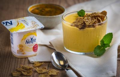 Fotografia em tons de amarelo em uma bancada de madeira, um paninho bege, um potinho com um creme de iogurte de maracujá com gengibre. Ao lado, um potinho de maracujá e uma colher.