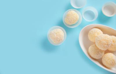 Fotografia vista de cima de cinco docinhos recheados com geleia de damasco e passados no coco dentro de um recipiente oval e fundo de cor branca. Ao lado tem quatro forminhas de brigadeiro, e duas estão com o docinho, e estão sobre uma mesa azul. Fotografia vista de cima de cinco docinhos recheados com geleia de damasco e passados no coco dentro de um recipiente oval e fundo de cor branca. Ao lado tem quatro forminhas de brigadeiro, e duas estão com o docinho, e estão sobre uma mesa azul.