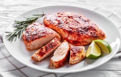 Fotografia em tons de branco com um prato branco e redondo ao centro. Em cima do prato existe dois filés de frango temperados com páprica. Um dos filés está cortado em varias fatias pequenas. Ao lado existe um ramo de alecrim e duas fatias de limão.