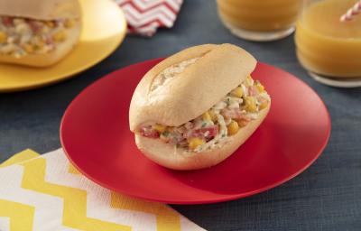 Foto da receita de sanduíche saudável sem glúten, montado em um pão francês recheado com um creme de frango, milho, sobre um prato vermelho e uma mesa com tecidos em tons amarelo e vermelho, ao lado de dois copos de suco e mais um sanduíche em outro prat Foto da receita de sanduíche saudável sem glúten, montado em um pão francês recheado com um creme de frango, milho, sobre um prato vermelho e uma mesa com tecidos em tons amarelo e vermelho, ao lado de dois copos de suco e mais um sanduíche em outro prat