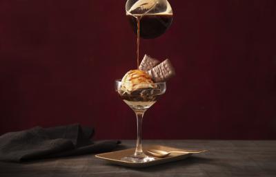 Fotografia em tons de preto de uma bancada de madeira com um prato quadrado, sobre ele uma taça com o affogato e uma jarra de café adicionando café á taça. Ao lado um paninho preto. Fotografia em tons de preto de uma bancada de madeira com um prato quadrado, sobre ele uma taça com o affogato e uma jarra de café adicionando café á taça. Ao lado um paninho preto.