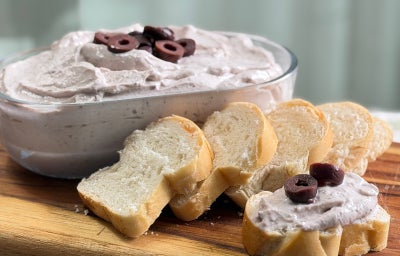 Fotografia mostra um pote transparente preenchido com maionese e rodelas de azeitona, ao lado fatias de pão.