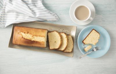 Fotografia em tons de azul em uma bancada de madeira clara com um prato redondo raso azul com uma fatia de bolo de Leite Moça. Ao lado, um recipiente retangular bege com o bolo inteiro. Fotografia em tons de azul em uma bancada de madeira clara com um prato redondo raso azul com uma fatia de bolo de Leite Moça. Ao lado, um recipiente retangular bege com o bolo inteiro.
