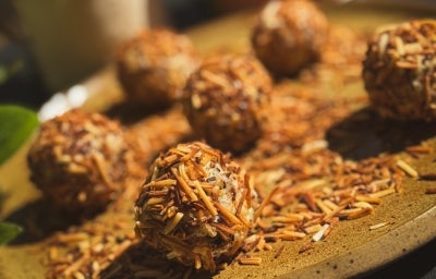 Fotografia em tons claros sob o sol de um prato grande, raso e de tom marrom claro. Sobre o prato tem alguns brigadeiros enrolados e passados no coco queimado, com mais coco queimado no prato. Ao fundo tem uma lata do leite ninho zero lactose.