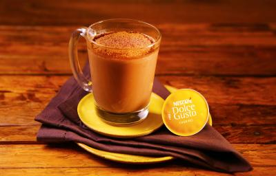 Fotografia em tons de amarelo em uma bancada de madeira escura, um prato amarelo redondo, um pano marrom e uma xícara de vidro com o chocafé com chocolate dentro dela.