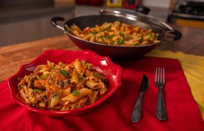 A fotografia mostra penne al dente coberto com molho de tomate caseiro, carne moída bem temperada, azeitonas e mostarda, finalizado com cebolinha e manjericão fresco.