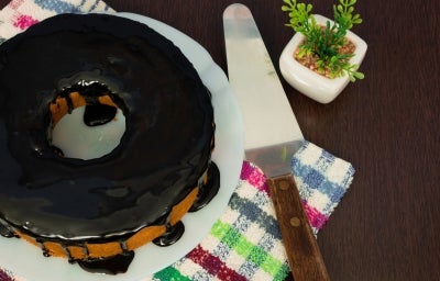 Um bolo de cenoura coberto com uma rica cobertura de chocolate, posicionado em um prato branco sobre uma superfície de madeira escura. Ao lado, um servidor de bolo de cabo de madeira e um pequeno vaso com uma planta artificial. Um bolo de cenoura coberto com uma rica cobertura de chocolate, posicionado em um prato branco sobre uma superfície de madeira escura. Ao lado, um servidor de bolo de cabo de madeira e um pequeno vaso com uma planta artificial.
