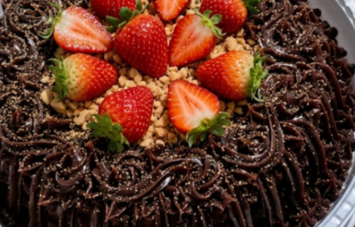 Bolo de chocolate com cobertura cremosa, morangos frescos e crocante granulado, sobre bandeja branca.