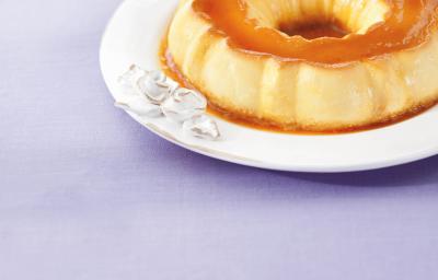 fotografia em tons de lilás, branco e laranja. Contém um prato redondo de branco com um pudim com calda de caramelo por cima. fotografia em tons de lilás, branco e laranja. Contém um prato redondo de branco com um pudim com calda de caramelo por cima.