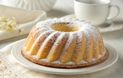 Um bolo tipo bundt (em formato de anel) com uma casquinha dourada e cobertura de açúcar de confeiteiro. Está servido em um prato escuro ao lado de uma xícara de café, criando um clima acolhedor e caseiro.