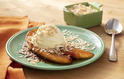 Fotografia em tons de branco em uma bancada de madeira de cor marrom. Ao centro, um prato redondo verde contendo 2 fatias de banana caramelizada com Neston e sorvete de creme. Ao fundo, uma colher, um recipiente contendo Neston e um pano laranja. Fotografia em tons de branco em uma bancada de madeira de cor marrom. Ao centro, um prato redondo verde contendo 2 fatias de banana caramelizada com Neston e sorvete de creme. Ao fundo, uma colher, um recipiente contendo Neston e um pano laranja.