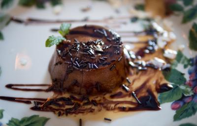 Fotografia de um mini pudim de chocolate sem furo no meio, decorado com granulado, calda de chocolate e uma folha de hortelã, sobre um prato branco desenhado.. Fotografia de um mini pudim de chocolate sem furo no meio, decorado com granulado, calda de chocolate e uma folha de hortelã, sobre um prato branco desenhado..