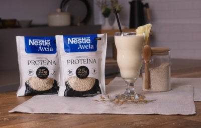 Foto da receita de shake pré-treino proteico servida em um copo de vidro alto sobre uma mesa de madeira com um pote de vidro cheio de aveia ao fundo