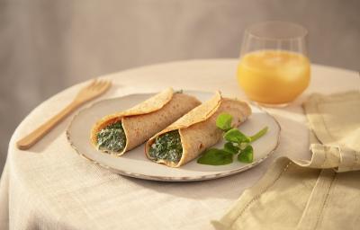 Foto em tons de bege da receita de panqueca de espinafre com ricota servida em duas porções sobre um prato de cerâmica claro em cima de uma mesa decorada com uma toalha bege, um copo de vidro com suco de laranja, um paninho verde e um garfo de madeira