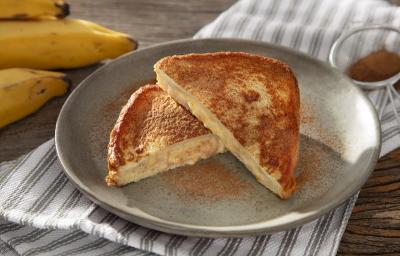 Foto em tons de cinza da receita de banana toast servida em duas metades sobre um prato de cerâmica cinza com canela em pó salpicada. Ao fundo há algumas bananas e um paninho listrado cinza