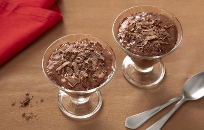 Fotografia de duas tacinhas de vidro ao lado de duas colheres de sopa. Dentro das tacinhas tem a mousse de nescau extra cacau com raspas de chocolate.