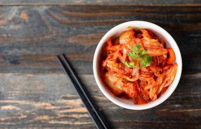 Fotografia vista de cima de um recipiente branco pequeno e fundo com uma receita de kimchi. Por cima da receita tem folhas de salsinha, e ao lado do recipiente tem dois hashis pretos sobre uma mesa de madeira escura. Fotografia vista de cima de um recipiente branco pequeno e fundo com uma receita de kimchi. Por cima da receita tem folhas de salsinha, e ao lado do recipiente tem dois hashis pretos sobre uma mesa de madeira escura.