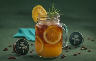 Fotografia em tons de verde de uma bancada verde, sobre ela um copo de vidro com cold brew com laranjas. Ao lado capsulas de Dolce Gusto Cold Brew e um paninho azul.