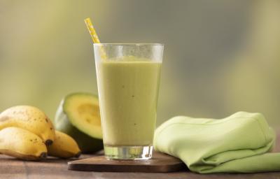 Fotografia em tons de verde e amarelo de uma bancada de madeira com um paninho verde e um apoio de copo de madeira, sobre ele um copo de vidro com a vitamina de abacate e um canudo amarelo. Ao fundo três bananas e meio abacate. Fotografia em tons de verde e amarelo de uma bancada de madeira com um paninho verde e um apoio de copo de madeira, sobre ele um copo de vidro com a vitamina de abacate e um canudo amarelo. Ao fundo três bananas e meio abacate.