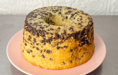Fotografia em tons claros com um bolo formigueiro ao centro. O bolo formigueiro é um bolo de massa clara feito com granulado de chocolate ao leite, o que traz uma aparência de vários pontinhos pretos ao bolo, similar a um formigueiro.
