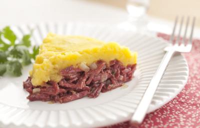 Fotografia em tons de vermelho em uma mesa de madeira clara com um prato redondo raso branco ao centro com uma fatia do escondidinho de carne seca com mandioquinha e coentro ao lado para enfeitar o prato. Fotografia em tons de vermelho em uma mesa de madeira clara com um prato redondo raso branco ao centro com uma fatia do escondidinho de carne seca com mandioquinha e coentro ao lado para enfeitar o prato.