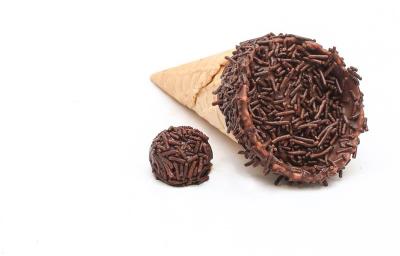 Fotografia de um cone trufado com brigadeiro sobre uma mesa off white. Ao lado do cone tem um brigadeiro passado no granulado.