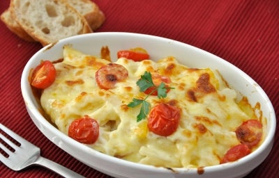 Em cima de uma mesa, refratário com batatas, queijo gratinado e tomates, decorado com folha de salsa.