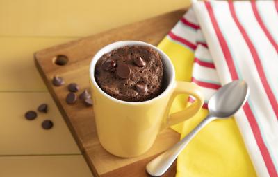 Fotografia em tons de amarelo em uma bancada de madeira amarela, um pano listrado vermelho e amarelo e uma tábua de madeira ao centro da foto com uma caneca amarela com o bolo de caneca de chocolate apoiada sobre ela. Fotografia em tons de amarelo em uma bancada de madeira amarela, um pano listrado vermelho e amarelo e uma tábua de madeira ao centro da foto com uma caneca amarela com o bolo de caneca de chocolate apoiada sobre ela.