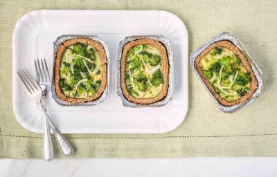 Fotografia em tons de branco, verde e dourado de uma bancada vista de cima mostrando refratário branco com duas porções de quiche com brócolis e dois garfos, com mais uma porção ao lado, tudo sobre guardanapo em tom de branco. Fotografia em tons de branco, verde e dourado de uma bancada vista de cima mostrando refratário branco com duas porções de quiche com brócolis e dois garfos, com mais uma porção ao lado, tudo sobre guardanapo em tom de branco.