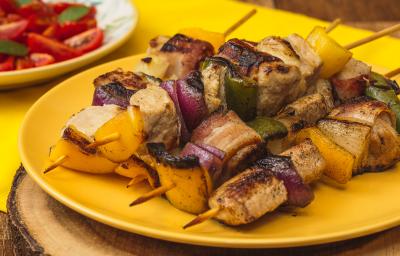 Foto bem aproximada de um prato amarelo sobre o qual há quatro espetinhos montados com carne de porco, cebolas roxa e tradicional e pimentões verdes e amarelos. Ao fundo há um prato branco com uma salada de tomates. Foto bem aproximada de um prato amarelo sobre o qual há quatro espetinhos montados com carne de porco, cebolas roxa e tradicional e pimentões verdes e amarelos. Ao fundo há um prato branco com uma salada de tomates.