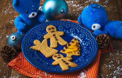 Fotografia em tons de laranja e azul em uma bancada de madeira, um paninho laranja, um prato azul com os biscoitinhos natalinos em cima dele. Ao lado, decoração e enfeites de natal. Fotografia em tons de laranja e azul em uma bancada de madeira, um paninho laranja, um prato azul com os biscoitinhos natalinos em cima dele. Ao lado, decoração e enfeites de natal.