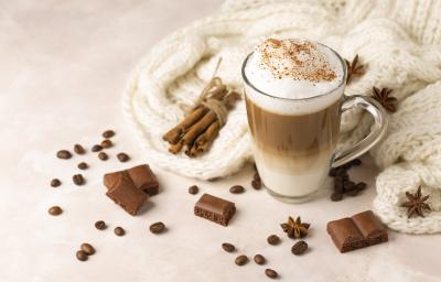 Fotografia em tons de branco de uma xícara de café de vidro com cappuccino feito de café, chocolate em pó, canela e laranja. Por cima do cappuccino tem canela salpicada, ao lado da xícara tem canelas em pau, chocolates, anis estrelado e uma lã branca.