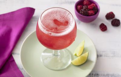Fotografia em tons de branco, roxo e verde de uma bancada de madeira branca e um prato redondo verde, sobre ele uma taça com o refresco de tônica e duas fatias de limão. Ao lado um paninho roxo e um recipiente redondo roxo com frutas vermelhas. Fotografia em tons de branco, roxo e verde de uma bancada de madeira branca e um prato redondo verde, sobre ele uma taça com o refresco de tônica e duas fatias de limão. Ao lado um paninho roxo e um recipiente redondo roxo com frutas vermelhas.