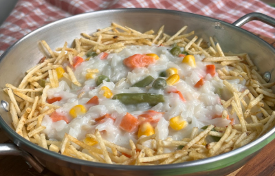 A imagem mostra uma panela de alumínio redonda com uma porção de arroz branco cremoso misturado com legumes picados e batata palha em volta. A panela está sobre um pano xadrez branco e vermelho.