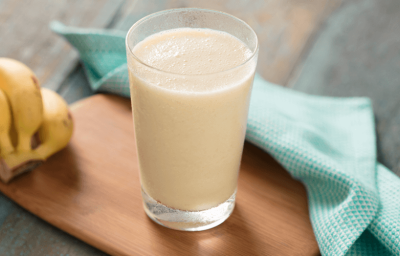 Fotografia em tons de azul e amarelo em uma bancada de madeira escura, uma tábua de madeira, um pano de pano azul com bolinhas, um cacho de bananas e um copo de vidro alto com a vitamina de banana com maracujá e coco. Fotografia em tons de azul e amarelo em uma bancada de madeira escura, uma tábua de madeira, um pano de pano azul com bolinhas, um cacho de bananas e um copo de vidro alto com a vitamina de banana com maracujá e coco.
