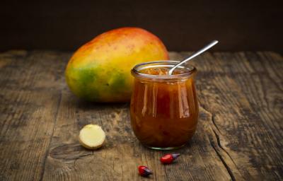 Fotografia de um chutney de manga e laranja temperado dentro de um recipiente de vidro pequeno, como se fosse um copo. Apoiado no chutney, uma colher de chá, e ao fundo, uma manga inteira com casca, sobre uma mesa de madeira. Fotografia de um chutney de manga e laranja temperado dentro de um recipiente de vidro pequeno, como se fosse um copo. Apoiado no chutney, uma colher de chá, e ao fundo, uma manga inteira com casca, sobre uma mesa de madeira.