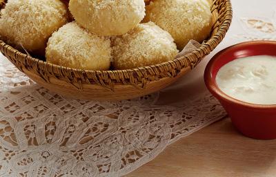 Fotografia em tons de branco, marrom, pardo e vermelho de uma bancada vista de cima. Um pano de renda branco e por cima um recipiente cor pardo e redondo com pães bolinha, ao lado um potinho vermelho redondo com creme branco. Fotografia em tons de branco, marrom, pardo e vermelho de uma bancada vista de cima. Um pano de renda branco e por cima um recipiente cor pardo e redondo com pães bolinha, ao lado um potinho vermelho redondo com creme branco.