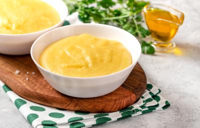 Na fotografia contém um pano branco com bolinhas verdes, por cima uma tábua de madeira de ponta redonda. Acima da tábua dois potes brancos com polenta dentro e ao lado um potinho com azeite. Na fotografia contém um pano branco com bolinhas verdes, por cima uma tábua de madeira de ponta redonda. Acima da tábua dois potes brancos com polenta dentro e ao lado um potinho com azeite.