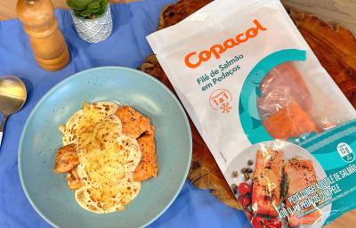 Fotografia em tons de azul e marrom com um prato azul e um pacote de salmão da marca Copacol ao centro. Dentro do prato existem dois filés de salmão cobertos com um molho de queijo gratinado.