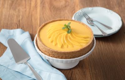 fotografia em tons de marrom, branco e amarelo de uma bancada marrom vista de frente, contém uma bailarina para servir sobremesa branca com uma torta por cima. Ao lado esquerdo contém um prato redondo branco e do lado direito um pano azul com uma espátula fotografia em tons de marrom, branco e amarelo de uma bancada marrom vista de frente, contém uma bailarina para servir sobremesa branca com uma torta por cima. Ao lado esquerdo contém um prato redondo branco e do lado direito um pano azul com uma espátula