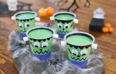 Imagem aproximada de três copos com a receita em tom verde, tudo decorado com tema de dia das bruxas, sobre uma bancada de madeira também decorada com teias falsas, aranhas e outros itens