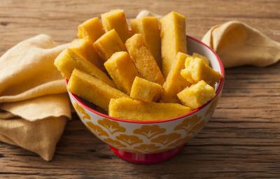 Fotografia em tons de amarelo de uma bancada de madeira com um pano amarelo retorcido e um potinho decorado branco, amarelo e vermelho, com vários palitos de polenta frita dentro. Fotografia em tons de amarelo de uma bancada de madeira com um pano amarelo retorcido e um potinho decorado branco, amarelo e vermelho, com vários palitos de polenta frita dentro.