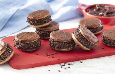 Fotografia em tons de vermelho em uma bancada de madeira clara, um pano azul, uma tábua vermelha com vários sanduíches de biscoito calipso recheados com brigadeiro de pão de mel. Ao fundo, um potinho vermelho com o brigadeiro dentro. Fotografia em tons de vermelho em uma bancada de madeira clara, um pano azul, uma tábua vermelha com vários sanduíches de biscoito calipso recheados com brigadeiro de pão de mel. Ao fundo, um potinho vermelho com o brigadeiro dentro.
