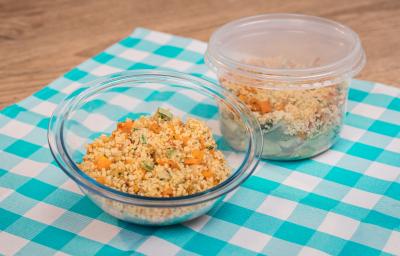 Foto de um pote de vidro redondo e um pote de plástico, ambos com a receita de salada de couscuz marroquino. Os dois estão sobre uma pano azul e branco quadriculado em uma bancada de madeira