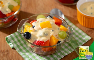 Salada de frutas coloridas em tigela de vidro, coberta com creme de maracujá, apoiada sobre guardanapo verde.